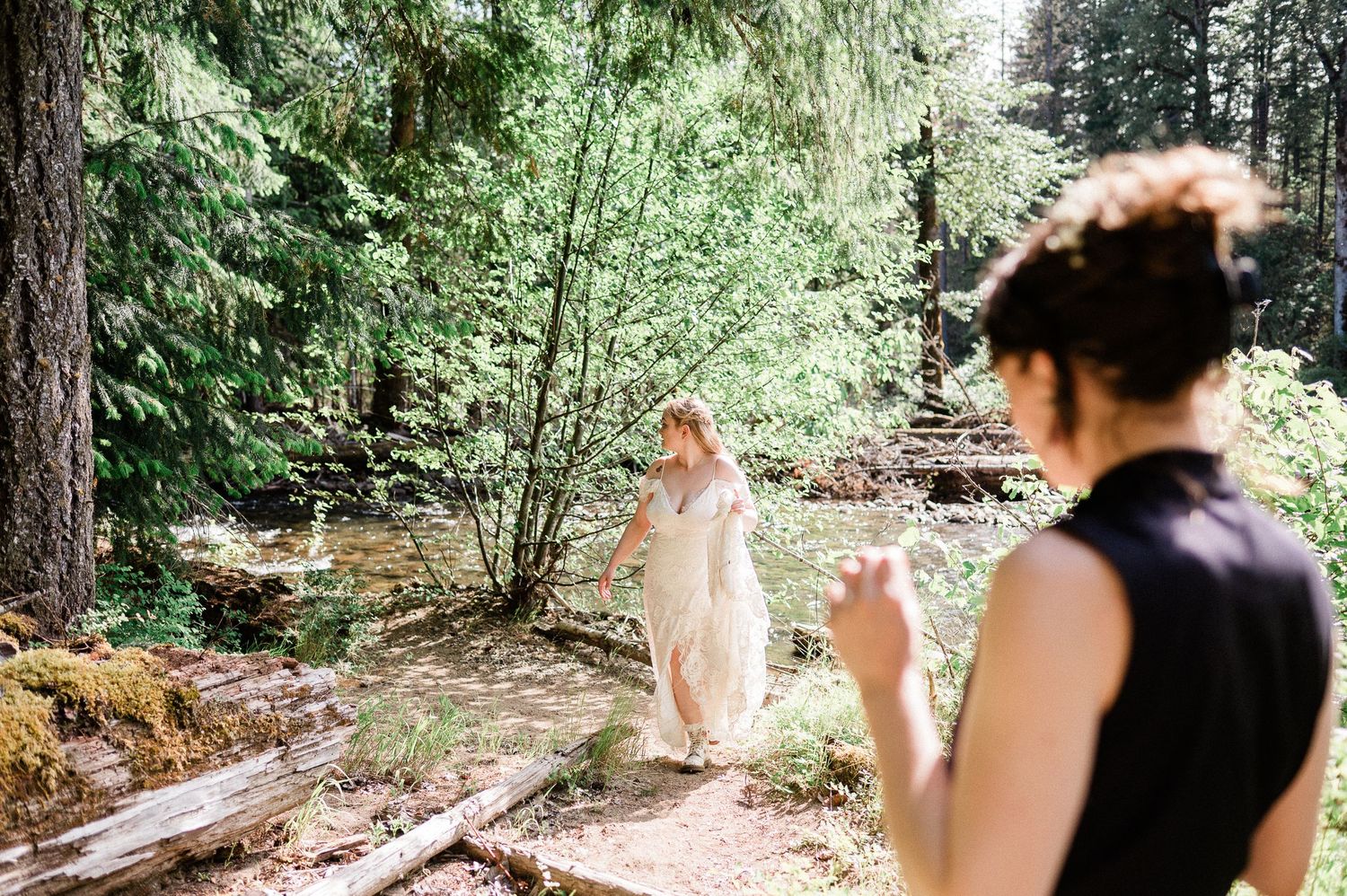A couple walks along a scenic forest trail surrounded by tall evergreen trees and dappled sunlight and a stream