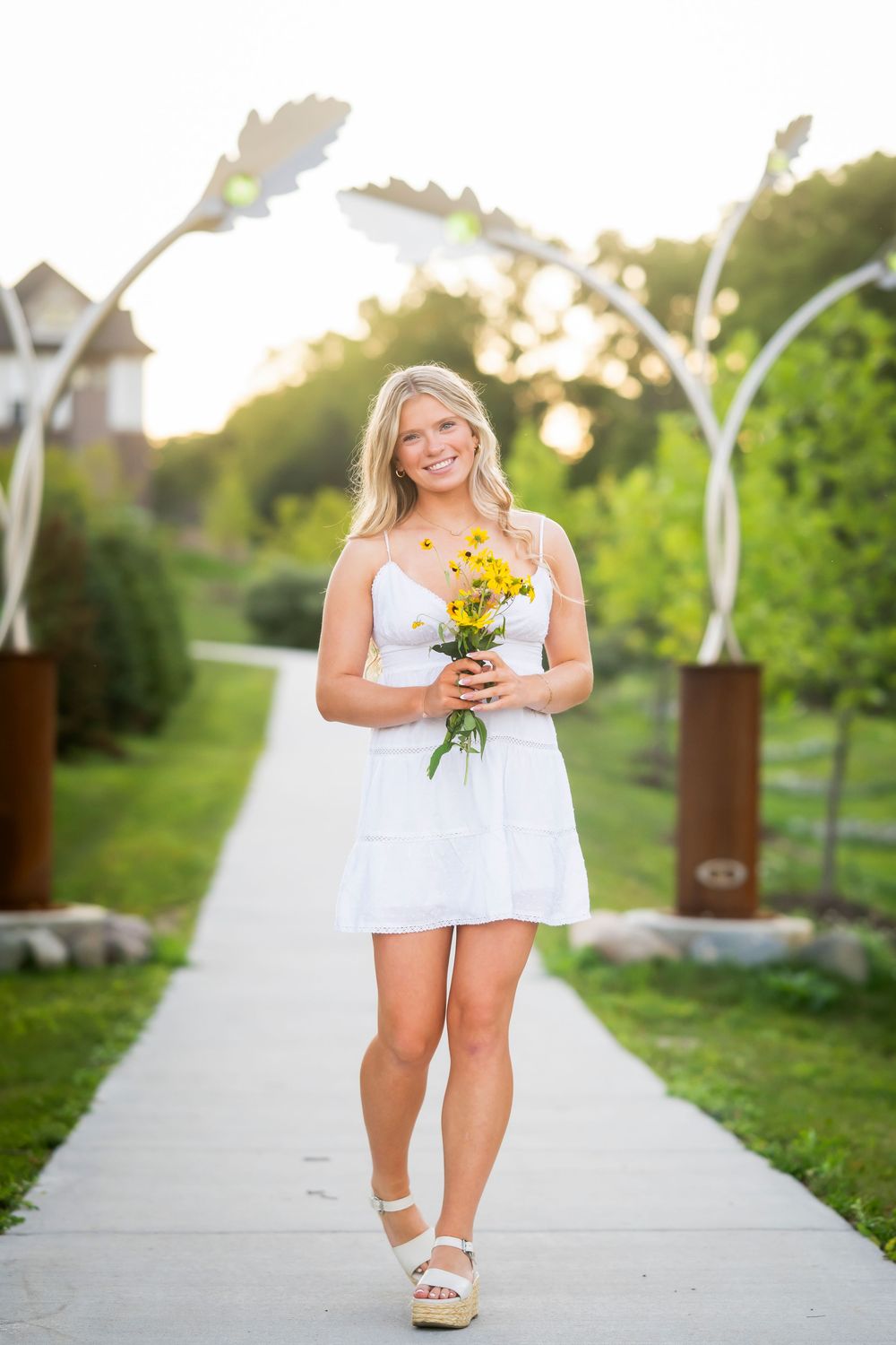 A person in a white sundress holds yellow flowers while walking along a garden path at sunset.