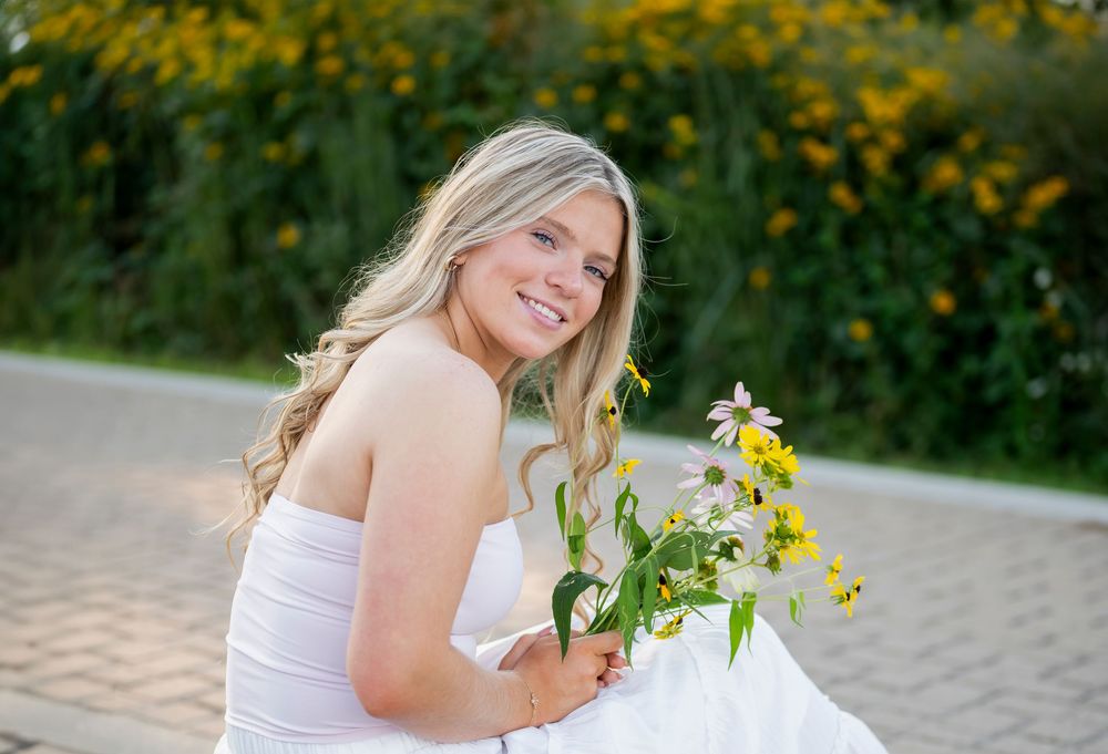 A person in a white dress holds yellow flowers while sitting on a path with wildflowers in the background.