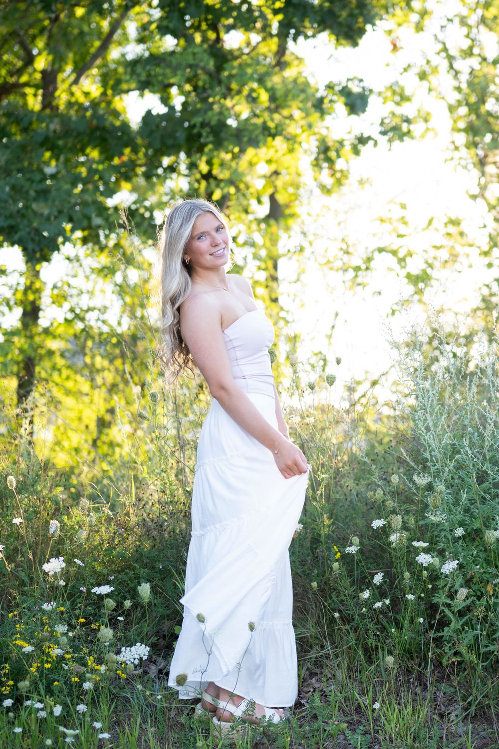 A person in a flowing white dress poses outdoors in a sunlit natural setting with wildflowers and trees.