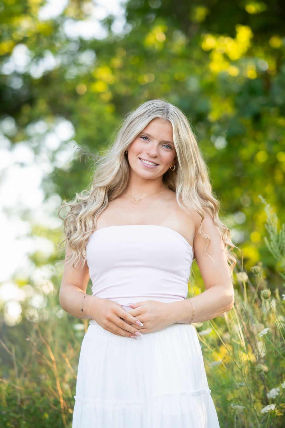 A woman in a white strapless dress poses outdoors in nature with soft sunlit greenery in the background.