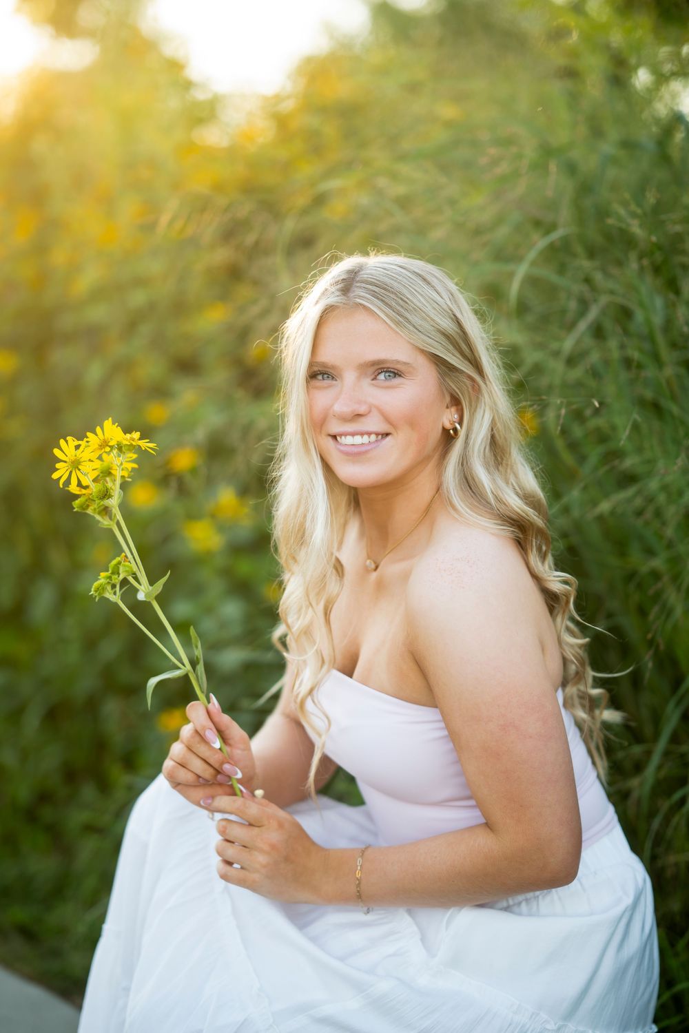 A person in a white dress holds a yellow wildflower while sitting outdoors during golden hour.