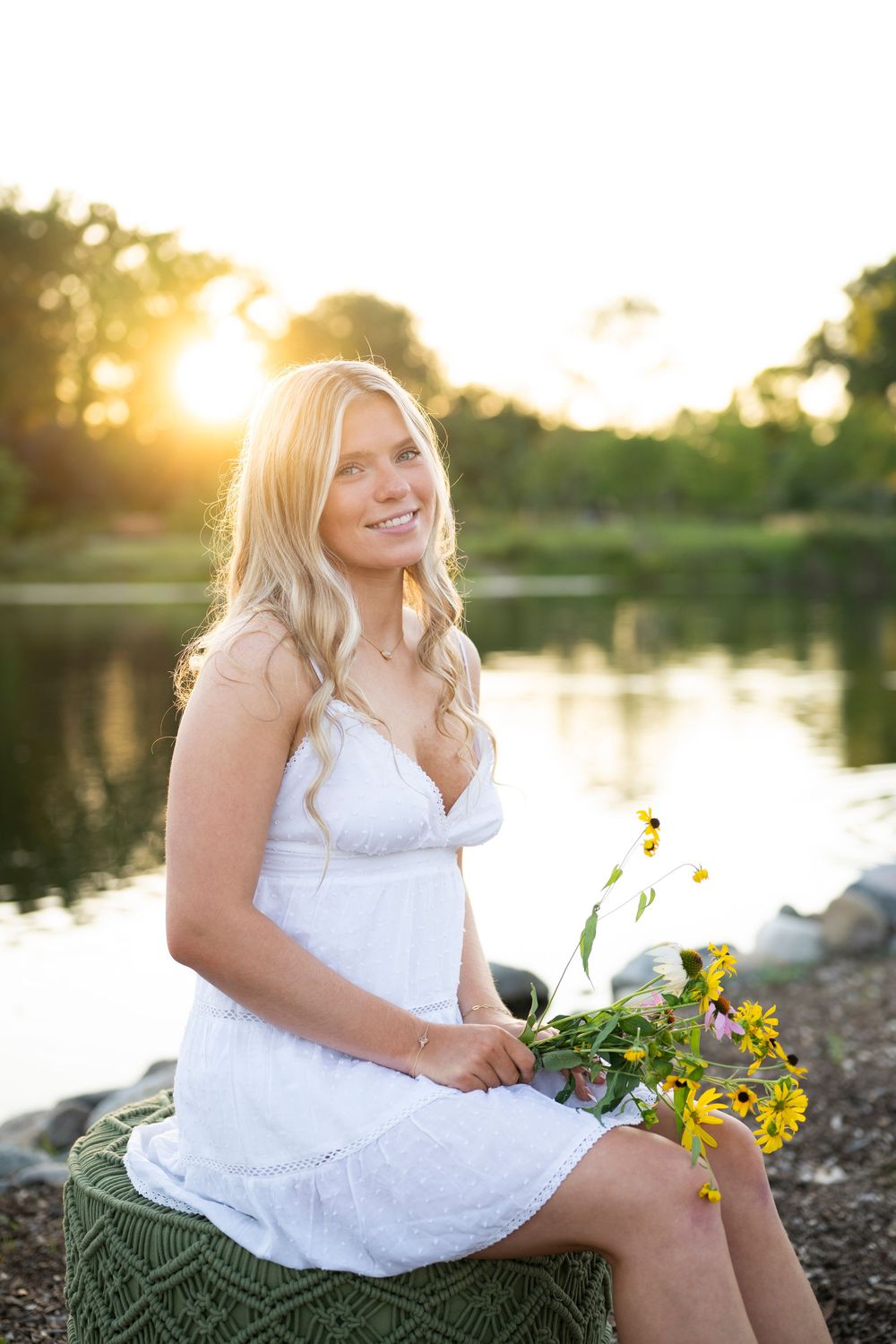 A person in a white dress sits by a lake at sunset holding yellow wildflowers in a dreamy outdoor portrait session.