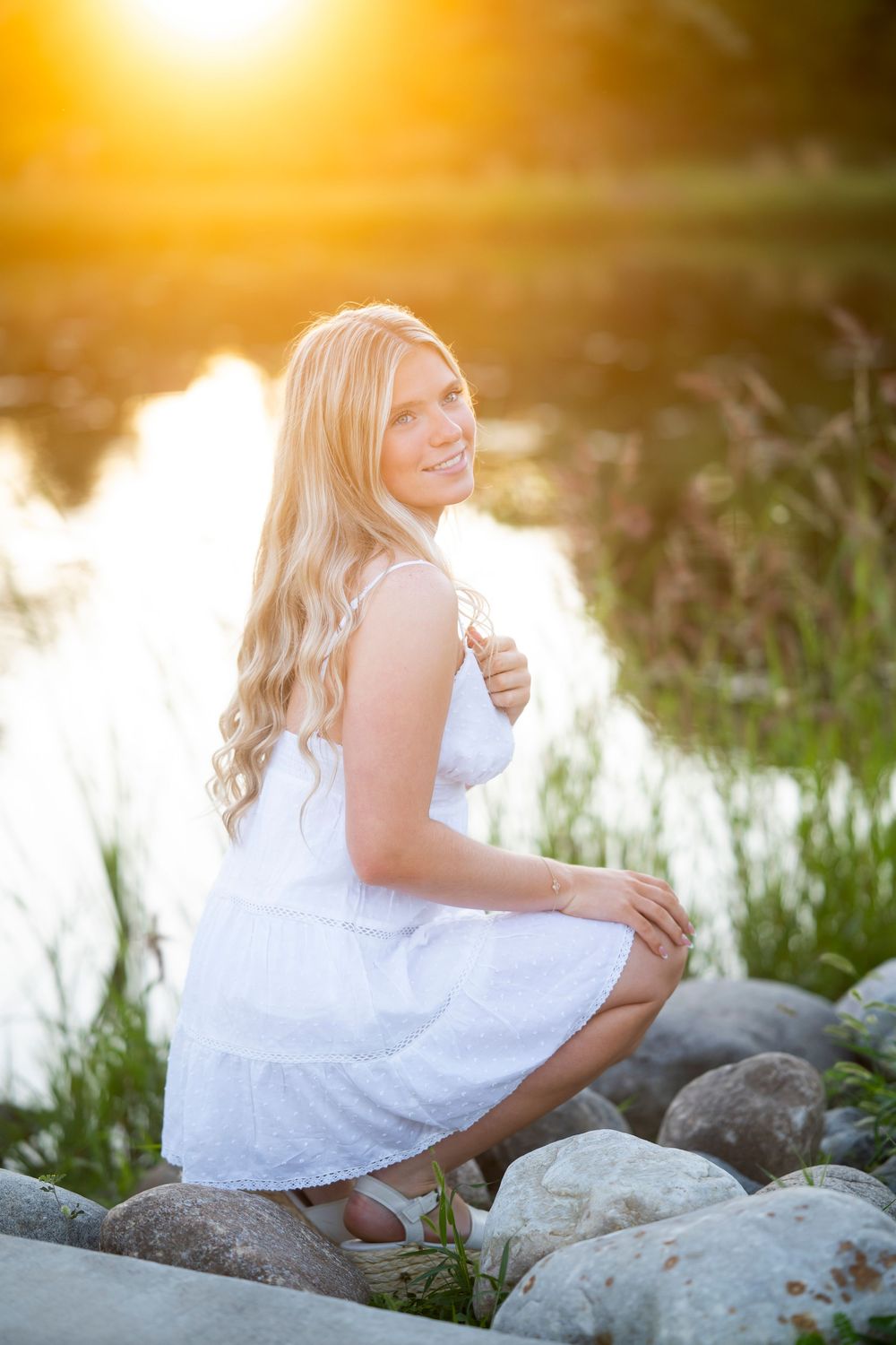 A person in a white dress poses on rocks by a lake at sunset with golden light creating a dreamy atmosphere.