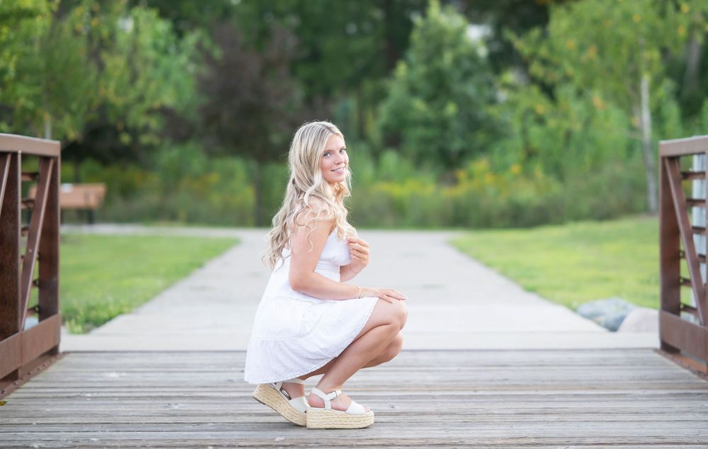 A person in a white dress sits thoughtfully on a wooden bridge in a serene outdoor setting.