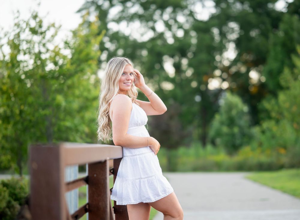 A person in a white dress poses by a wooden railing on a garden path during sunset.
