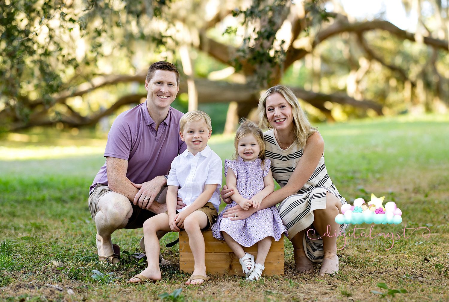 A family poses together outdoors on a sunny day under oak trees with Spanish moss in a park setting.