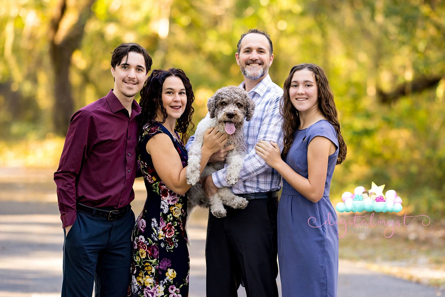 A group stands together on a wooded path during autumn while holding a small fluffy dog in the middle.