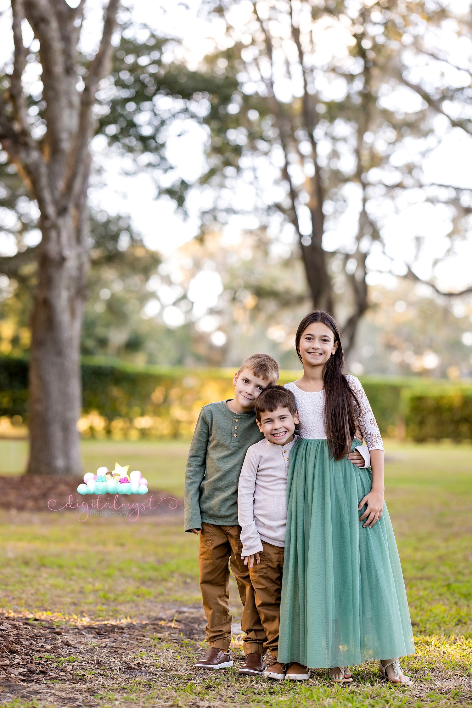 A family in coordinating green and neutral outfits poses together outdoors in a sunlit wooded setting.