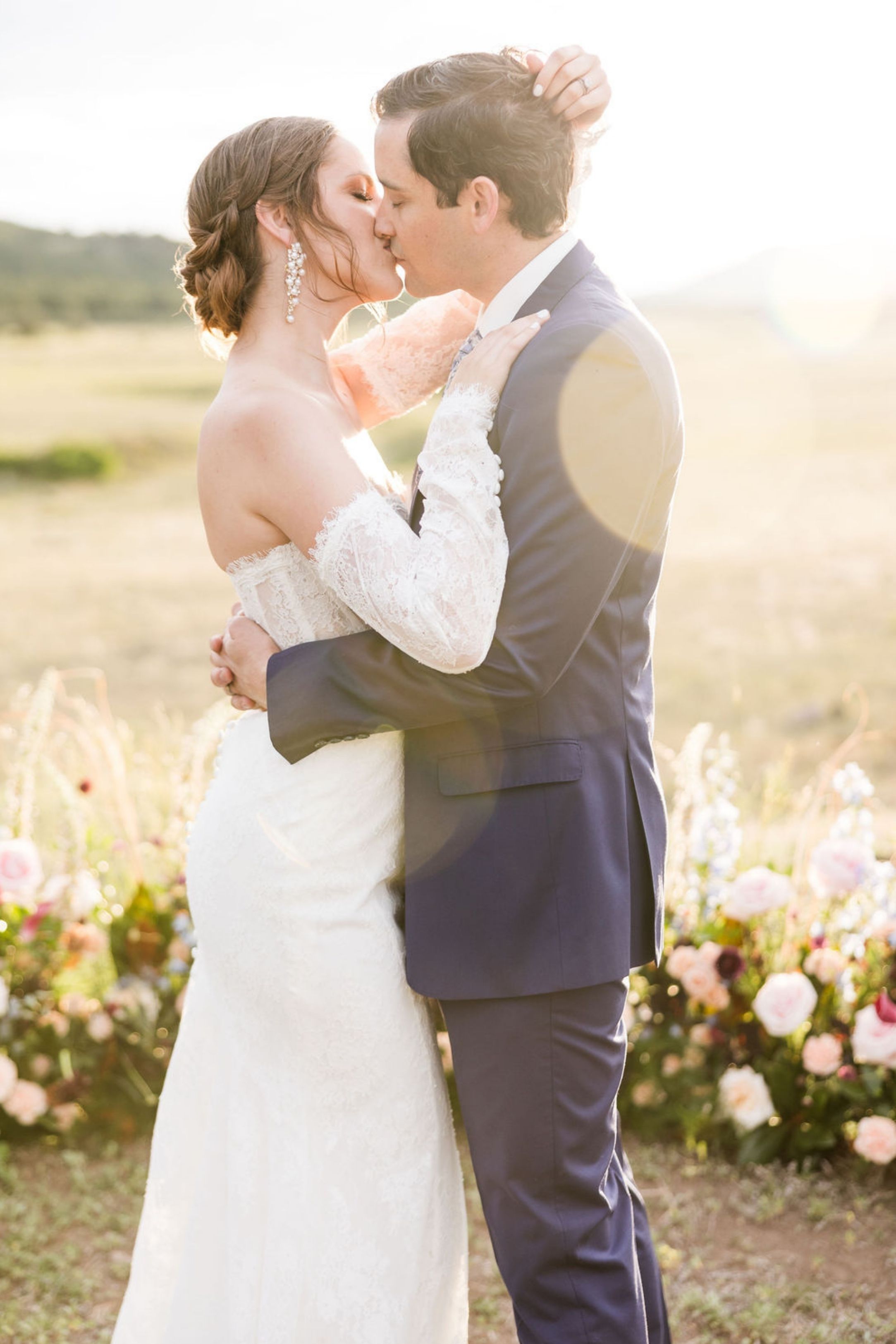 Bride and groom share first kiss at outdoor wedding ceremony surrounded by pink florals in golden sunlight.