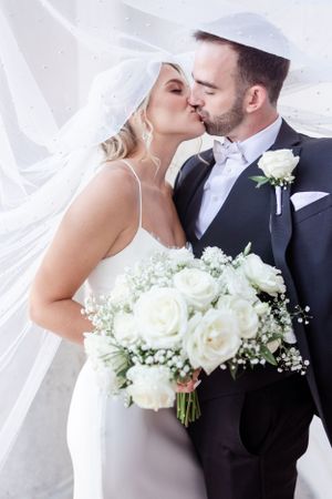Newlyweds share a romantic kiss under flowing veil while holding white rose bouquet.