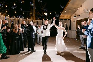 Newlyweds make grand entrance at outdoor wedding reception with bubbles and cheering guests under string lights.