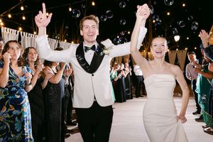 Bride and groom celebrate joyfully at wedding reception with raised arms, bubbles floating around them, guests cheering in background.