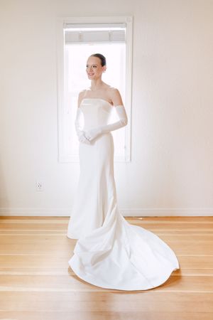 Elegant bride in a strapless white satin gown with long gloves and train, standing by a bright window on hardwood floors.