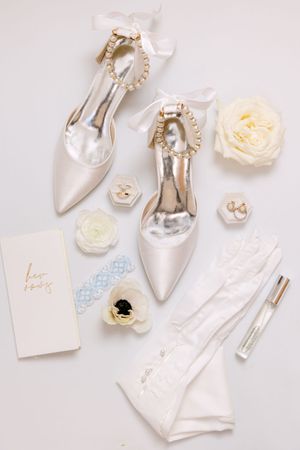 Elegant bridal flat lay with pearl-adorned satin heels, white roses, lace gloves, blue garter, and gold jewelry on white background.