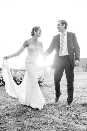 Black and white photo of a smiling bride and groom holding hands and walking outdoors on their wedding day.