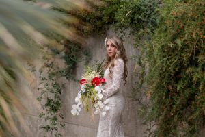 A bride in a white dress poses against a rustic stone wall surrounded by lush greenery and red flowers.