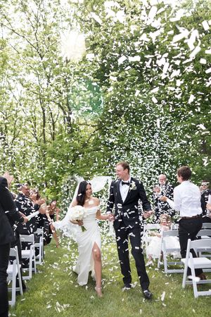 Bride and groom walk down outdoor aisle as guests toss white confetti petals during sunny garden wedding ceremony.