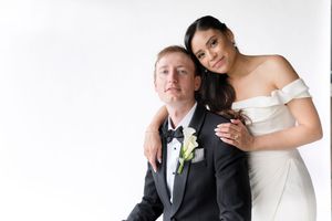 Bride in off-shoulder white gown embraces groom in black tuxedo with white boutonniere against a white studio backdrop.