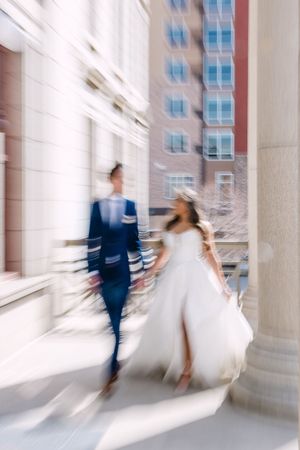 Bride and groom walking hand in hand along a sunlit urban colonnade, captured with a dreamy motion blur effect.