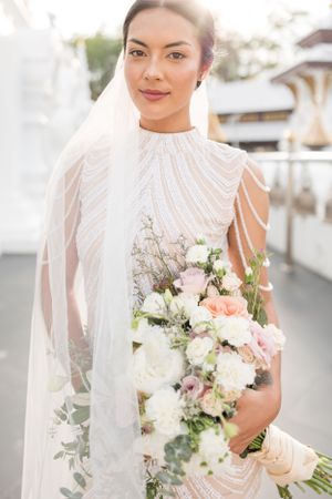 Bride in elegant beaded gown holds delicate bouquet of pink and white flowers with trailing greenery.