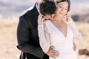 A groom in a black suit tenderly kisses his bride's neck as she smiles in a white lace wedding dress outdoors.