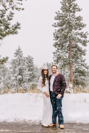 Couple poses in snowy winter wonderland, bride in lace gown with cowboy hat, groom in burgundy blazer.