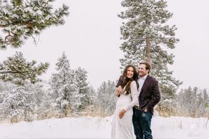 Couple embracing in snowy winter forest with snow-covered pine trees, bride in white gown and groom in burgundy blazer.