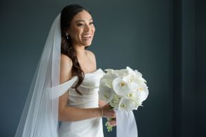 A bride wearing an off-shoulder wedding dress holds a white orchid bouquet against a dark backdrop.
