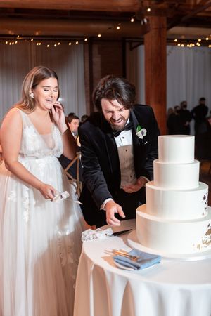 Bride and groom smiling while cutting their white four-tier wedding cake at their elegant reception venue.