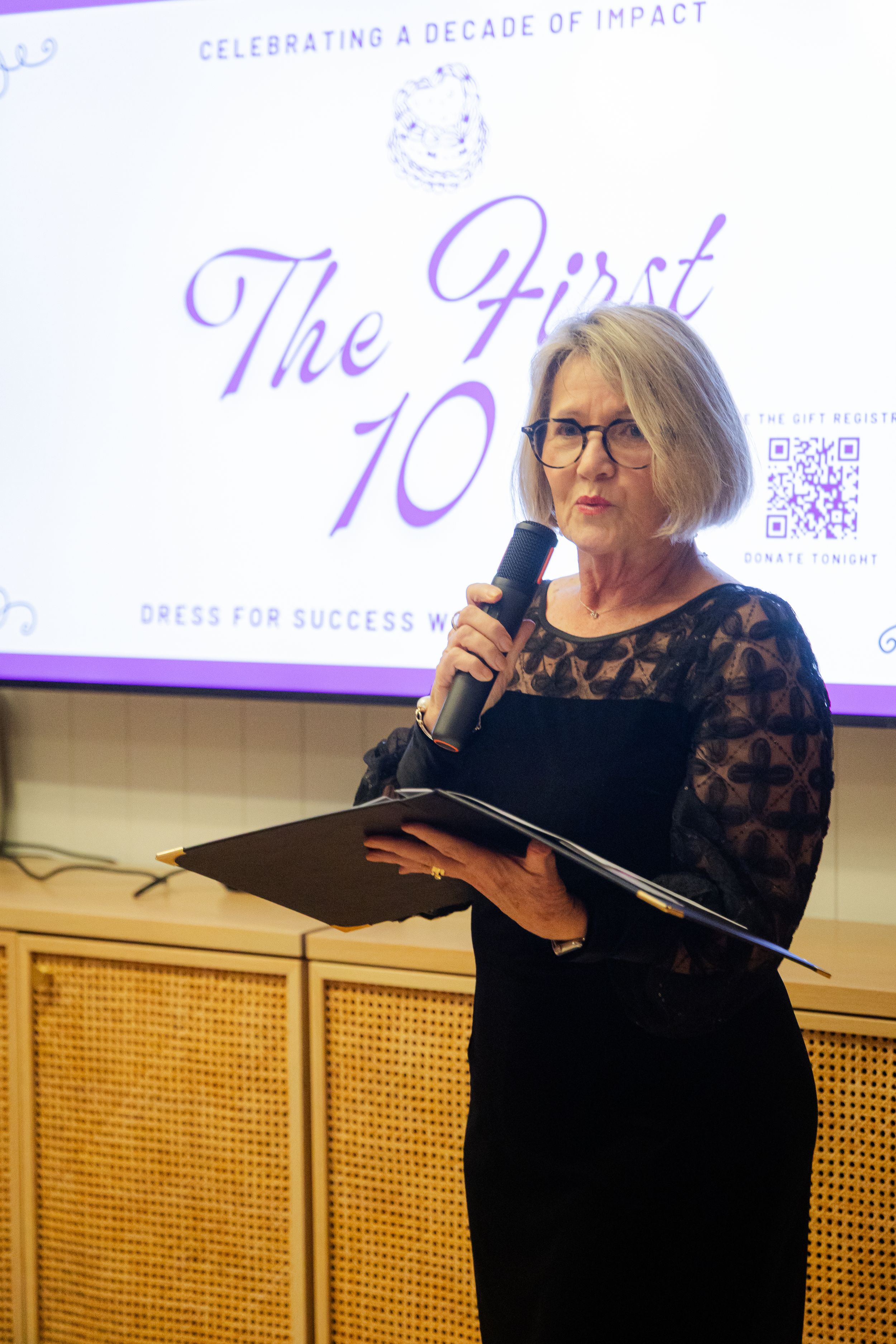 Woman in black lace dress speaks at Dress for Success event celebrating a decade of impact.