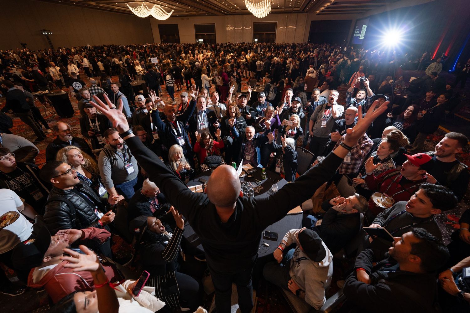 Steve J Larsen raises arms before a large crowd in a packed conference hall with bright off camera light capturing emotion and energy.