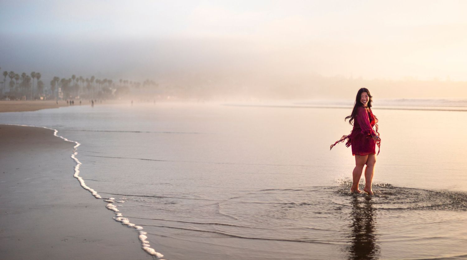Woman in red dress strolling along a foggy beach at sunset with gentle waves.
