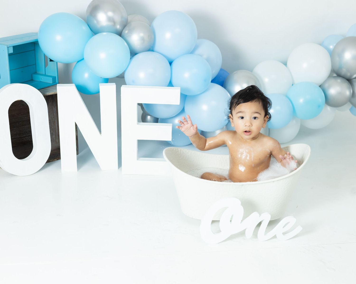 A baby sits in a bathtub surrounded by blue and white balloons for a first birthday photoshoot.