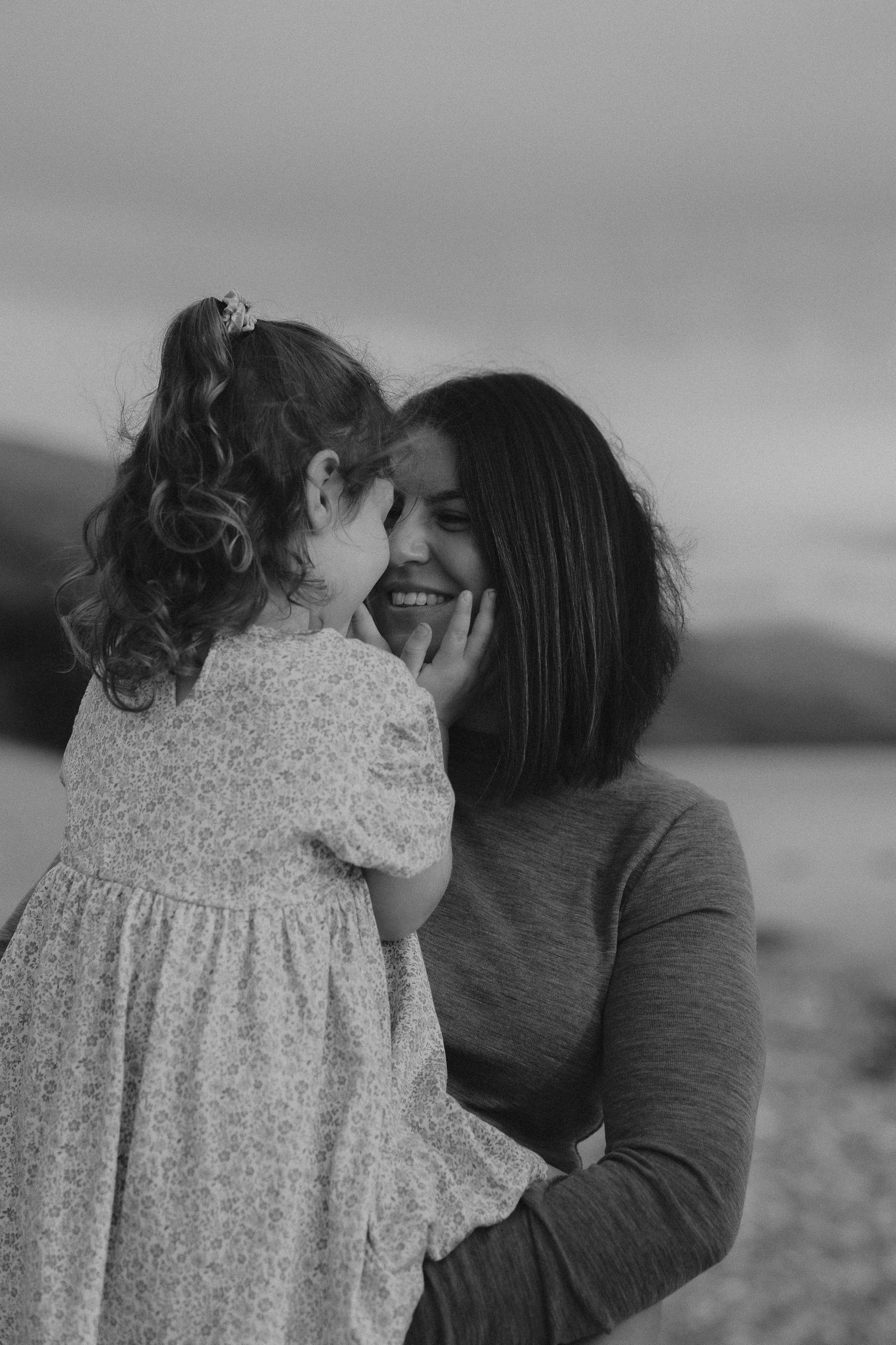 Black and white photo of a smiling mother holding her young daughter close, sharing a tender moment outdoors.