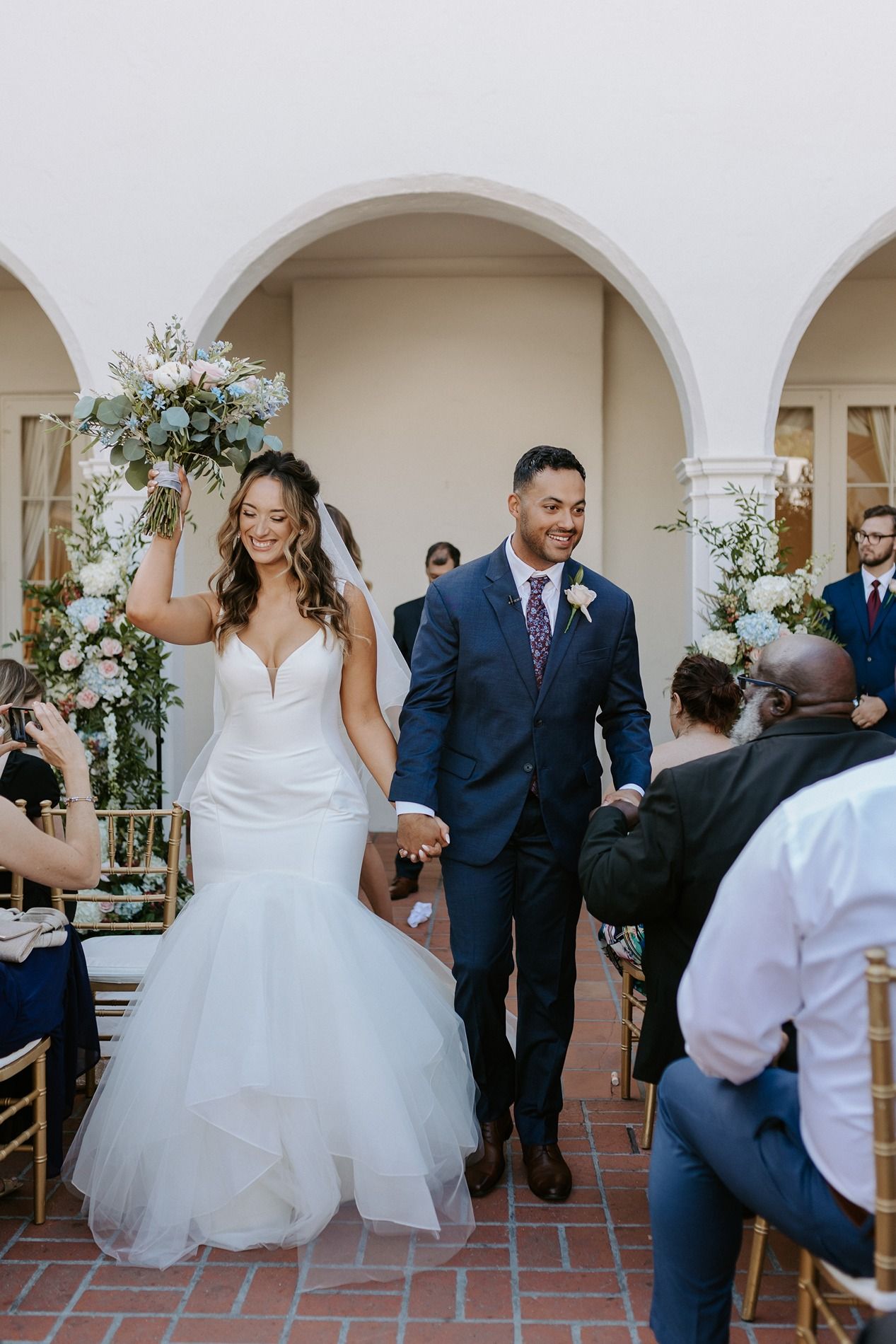 Bride in mermaid gown raises bouquet while walking down aisle with groom in navy suit at outdoor wedding ceremony at Darlington House.