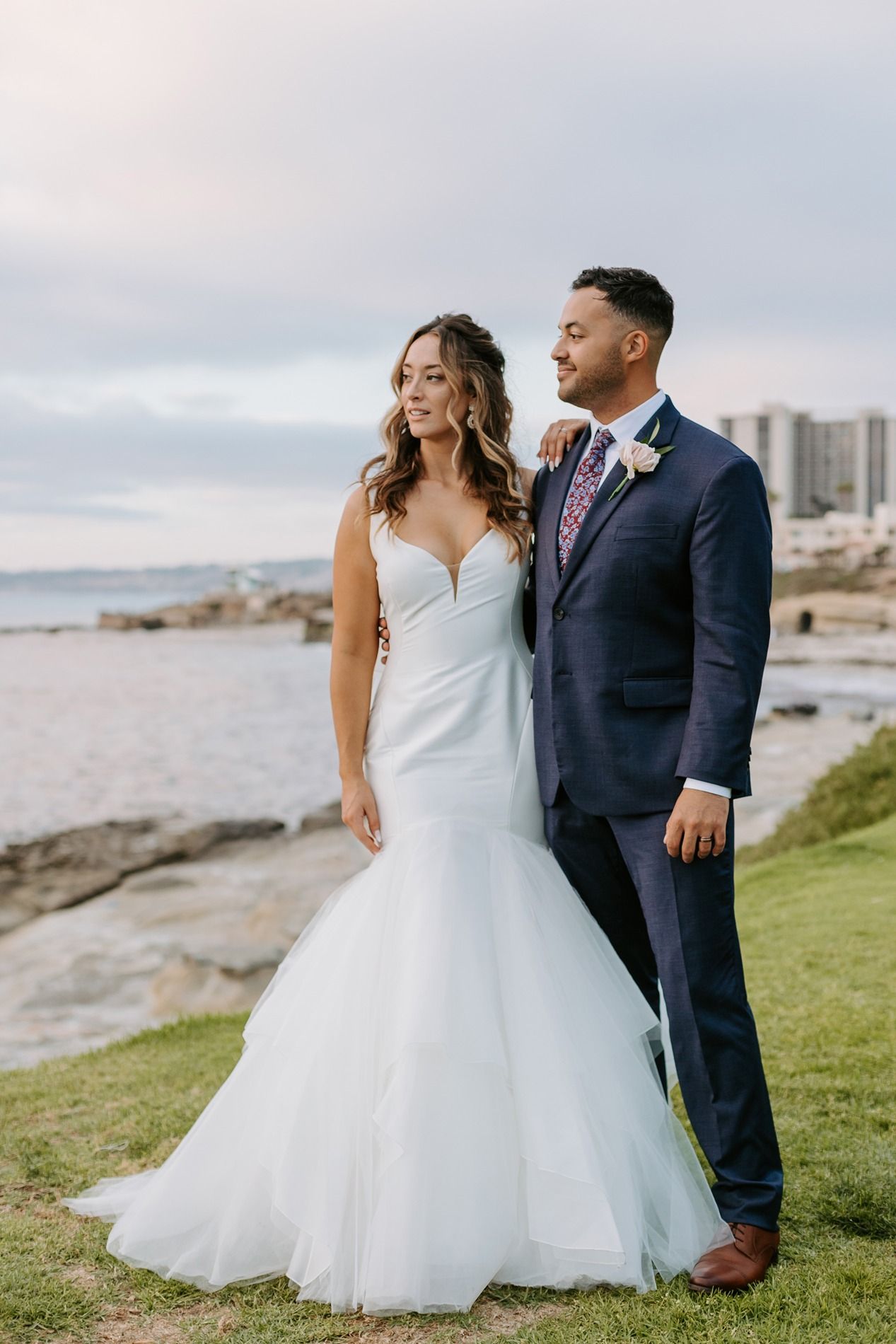 Bride in white mermaid gown and groom in navy suit pose together by the ocean on their wedding day in La Jolla.