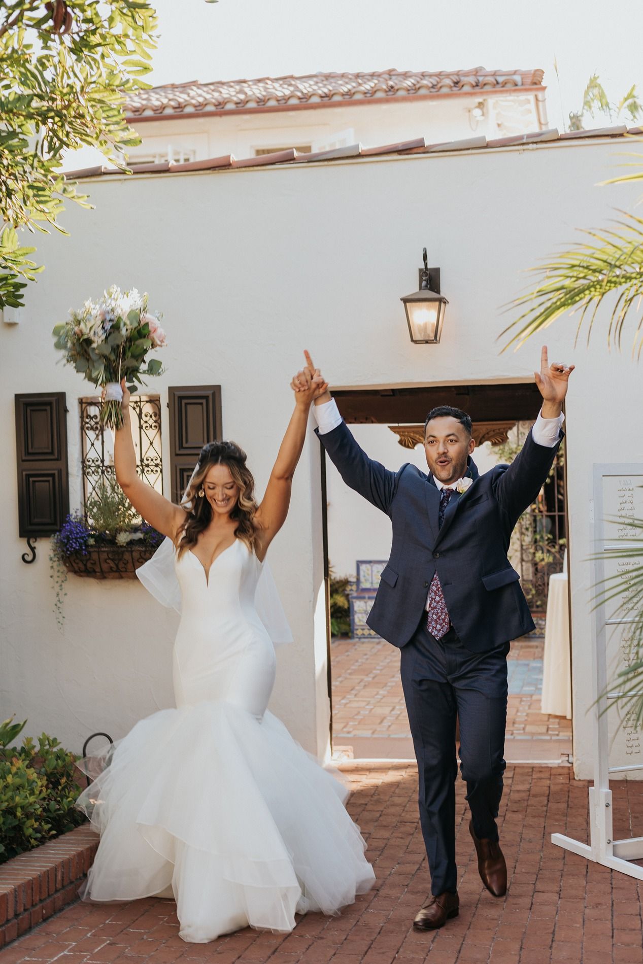 Joyful newlyweds celebrate their wedding exit, bride in mermaid gown holding bouquet, groom in navy suit, arms raised at Darlington House.