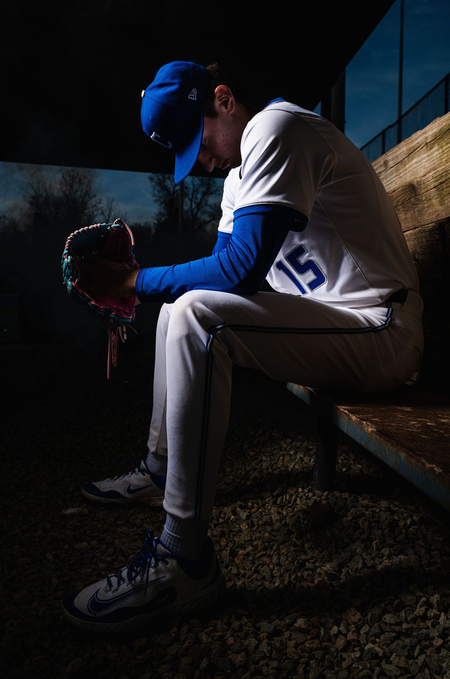 Baseball player in blue uniform sitting in dugout holding bat with glove and ball nearby.