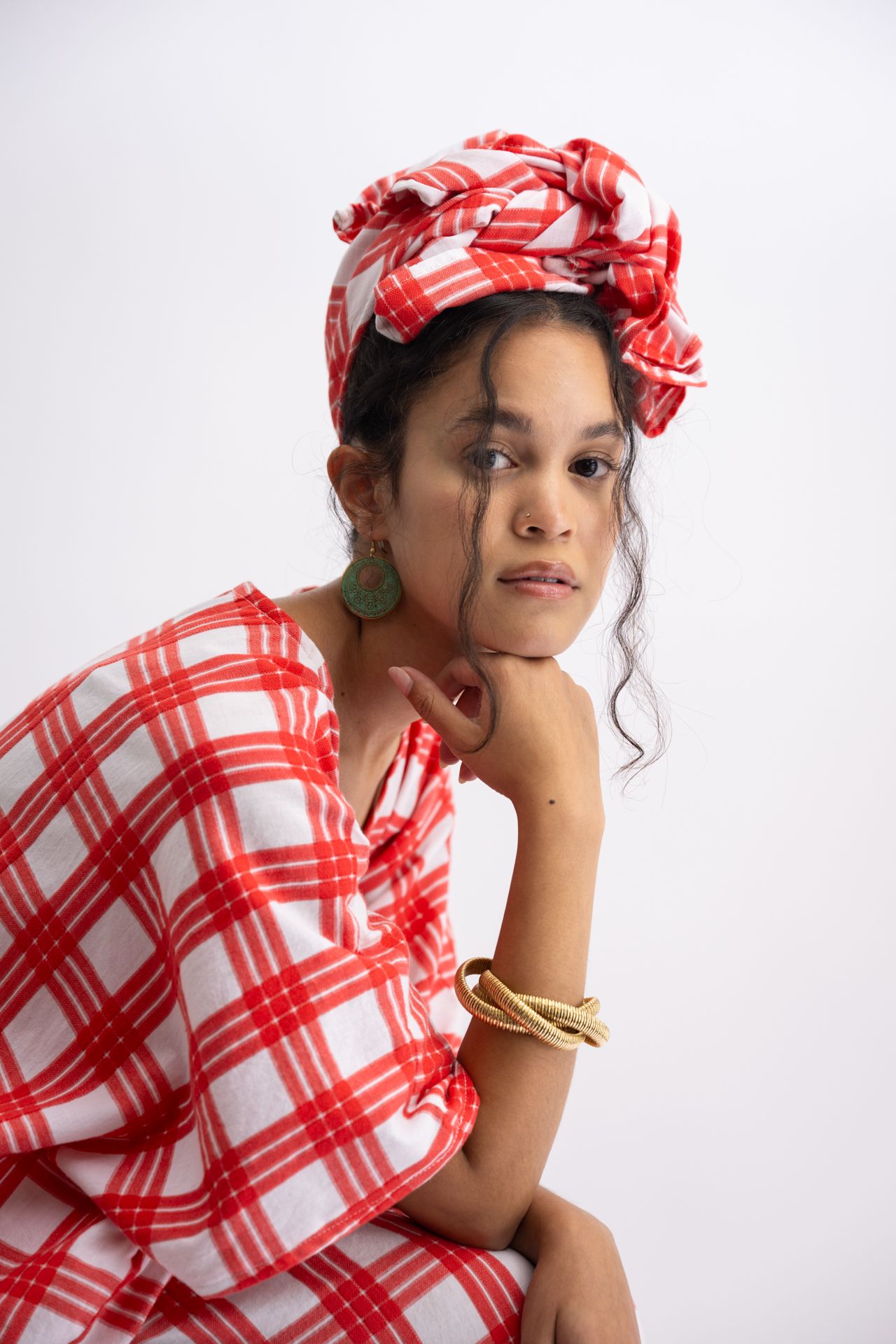 Woman in red plaid wrap dress and matching head wrap, gold bangles, green earrings, posing against white background.