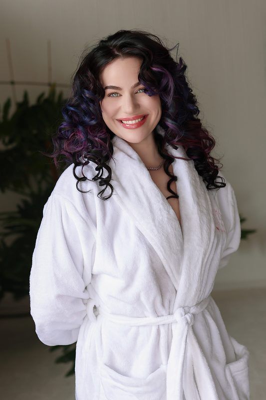 A person wearing a white bathrobe smiles while showing off curly purple and blue ombre hair.