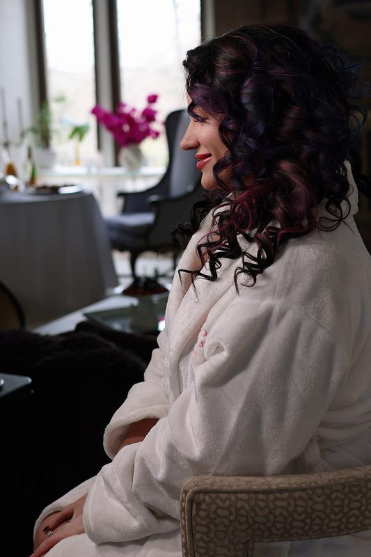 A person with dark curly hair wearing a white robe sits in profile at a salon with pink flowers in the background.