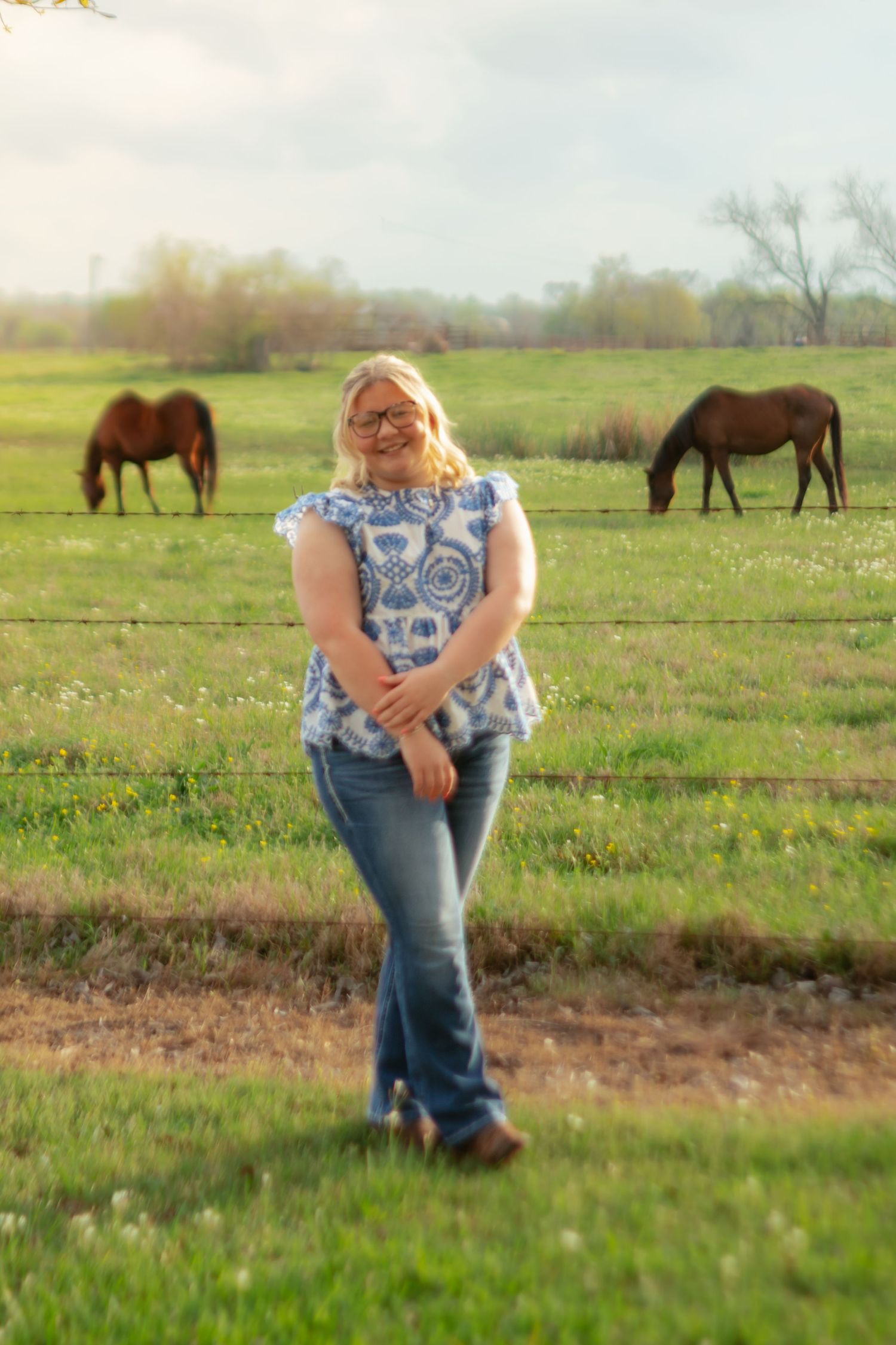 A figure stands in a grassy field with grazing horses visible in the background on a sunny spring day.