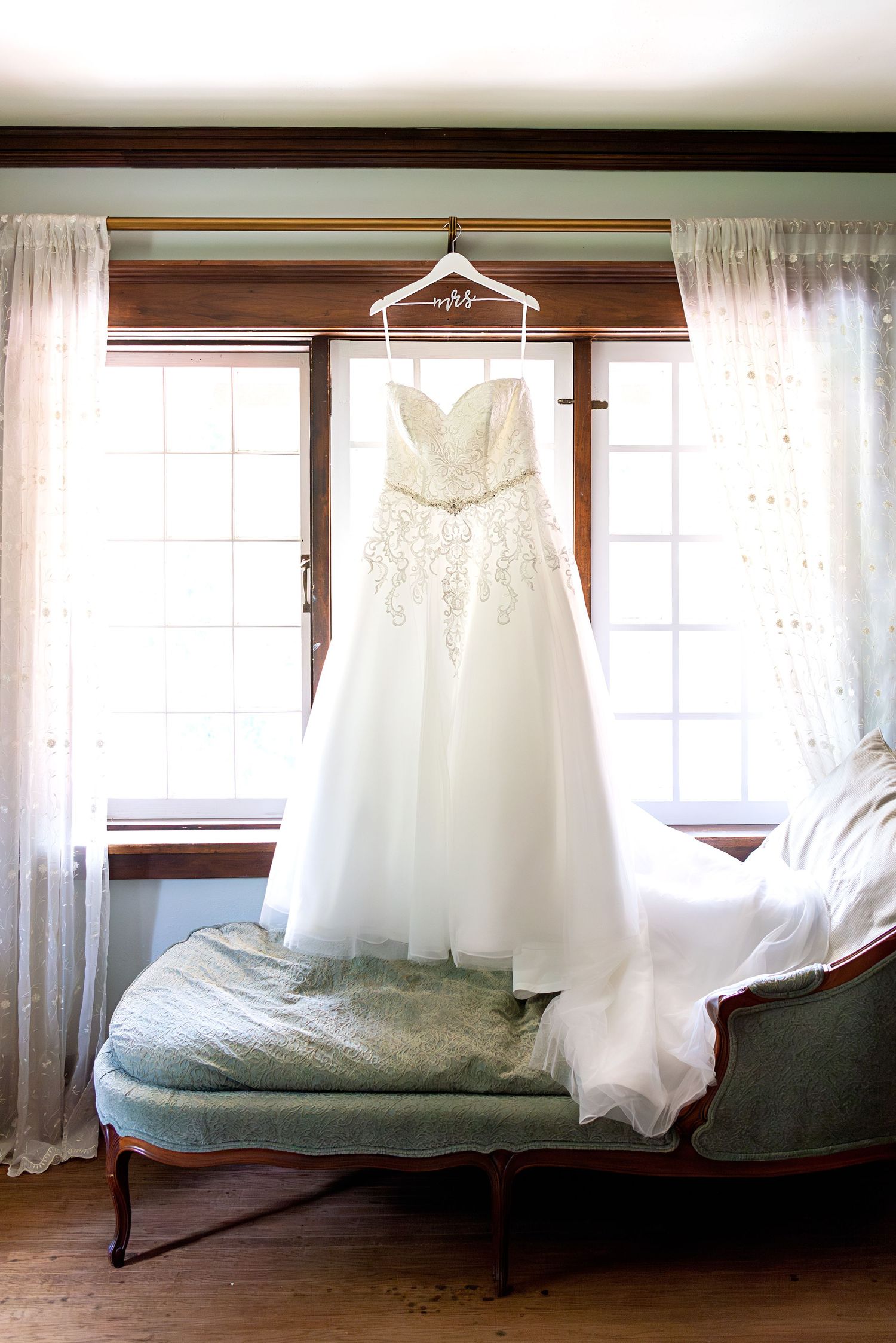 Elegant strapless A-line wedding gown with lace bodice hanging on a personalized hanger in a window above a vintage chaise lounge.