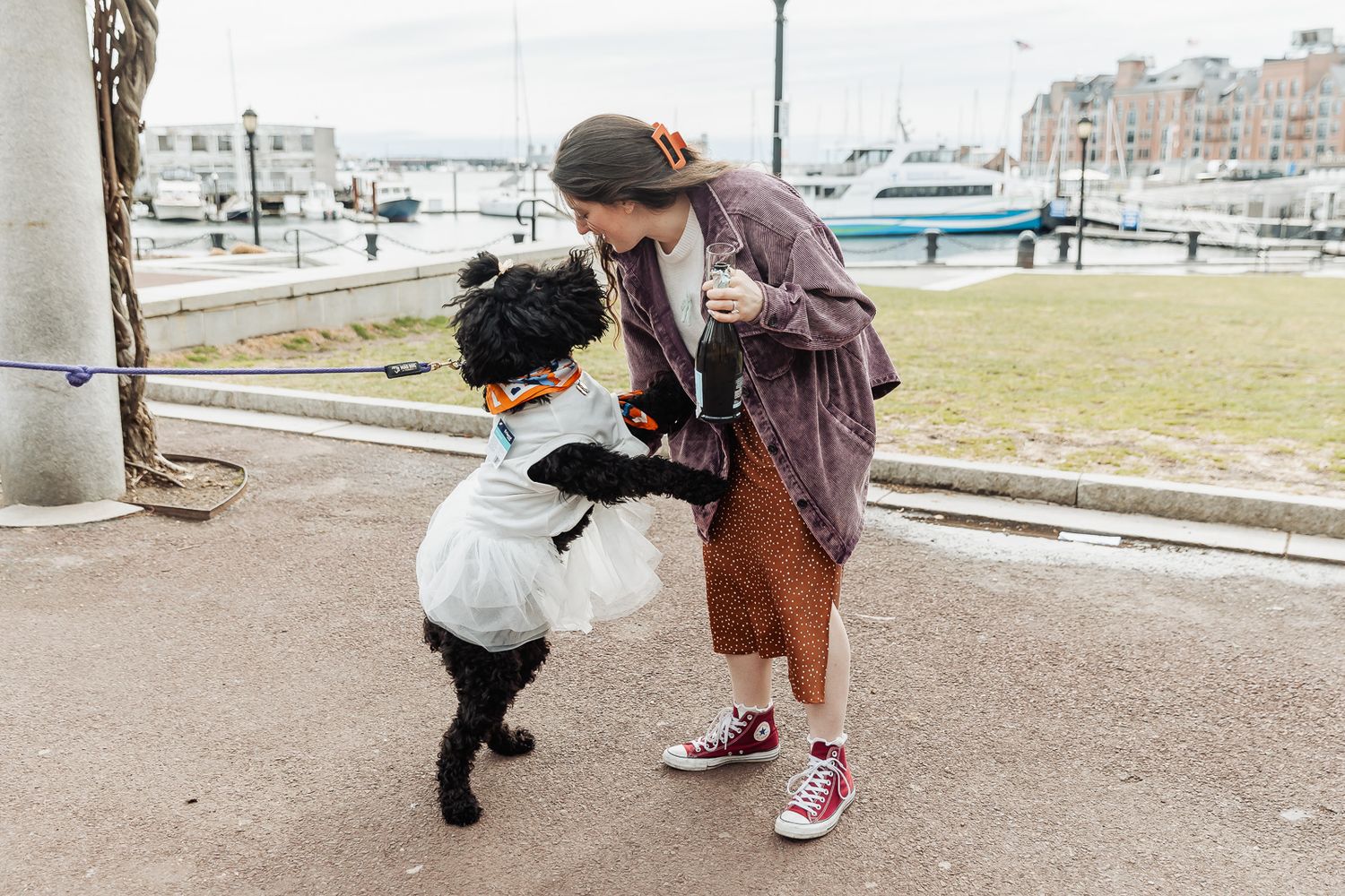 A woman plays with her black dog dressed in a ghost Halloween costume near a harbor waterfront.