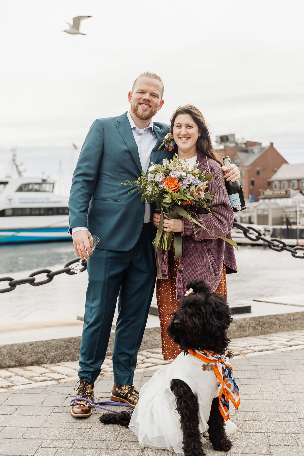 Couple in wedding attire pose with black dog by waterfront, woman holding colorful bouquet, man in teal suit.