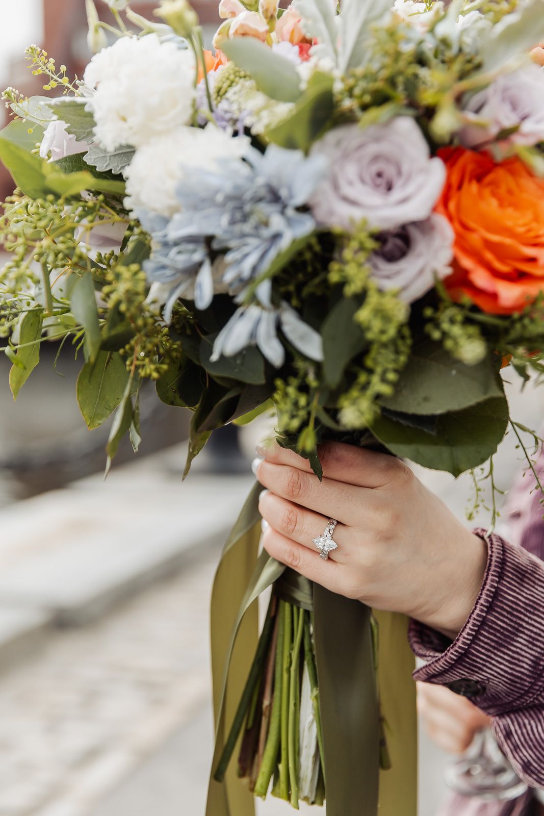 Bride holding colorful wedding bouquet with orange, white, and purple flowers, wearing a diamond engagement ring.