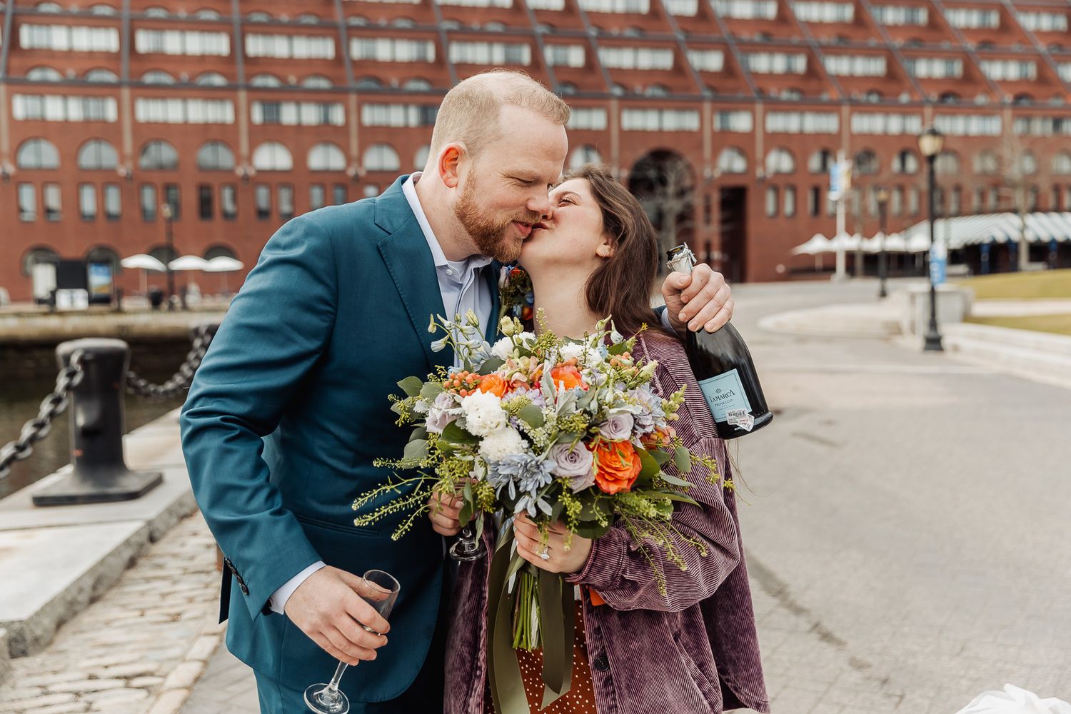 A couple kisses while holding a bouquet and champagne bottle outside a brick building.