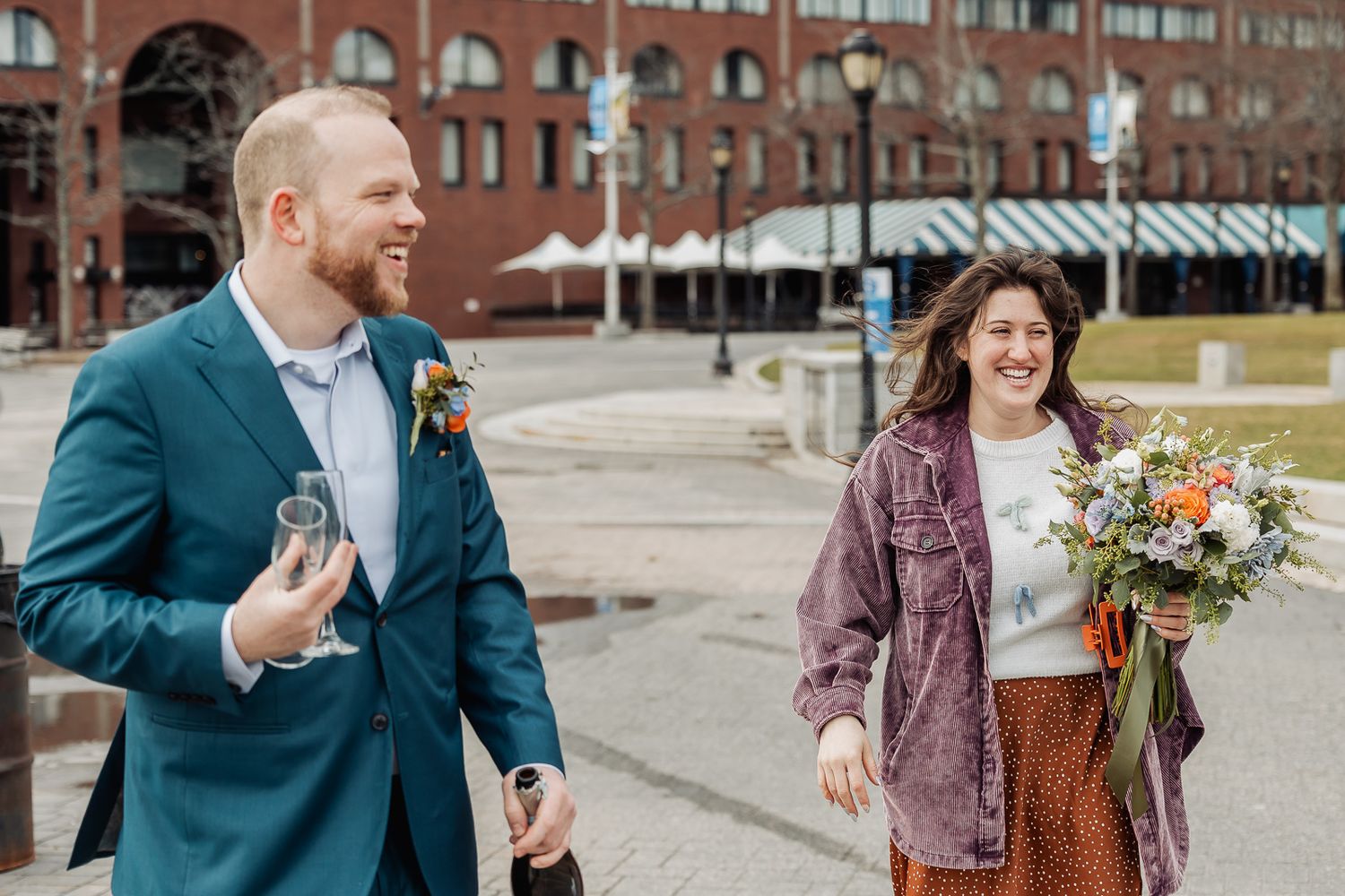 Couple walking outdoors after wedding ceremony, groom in blue suit with boutonniere, bride holding colorful bouquet.