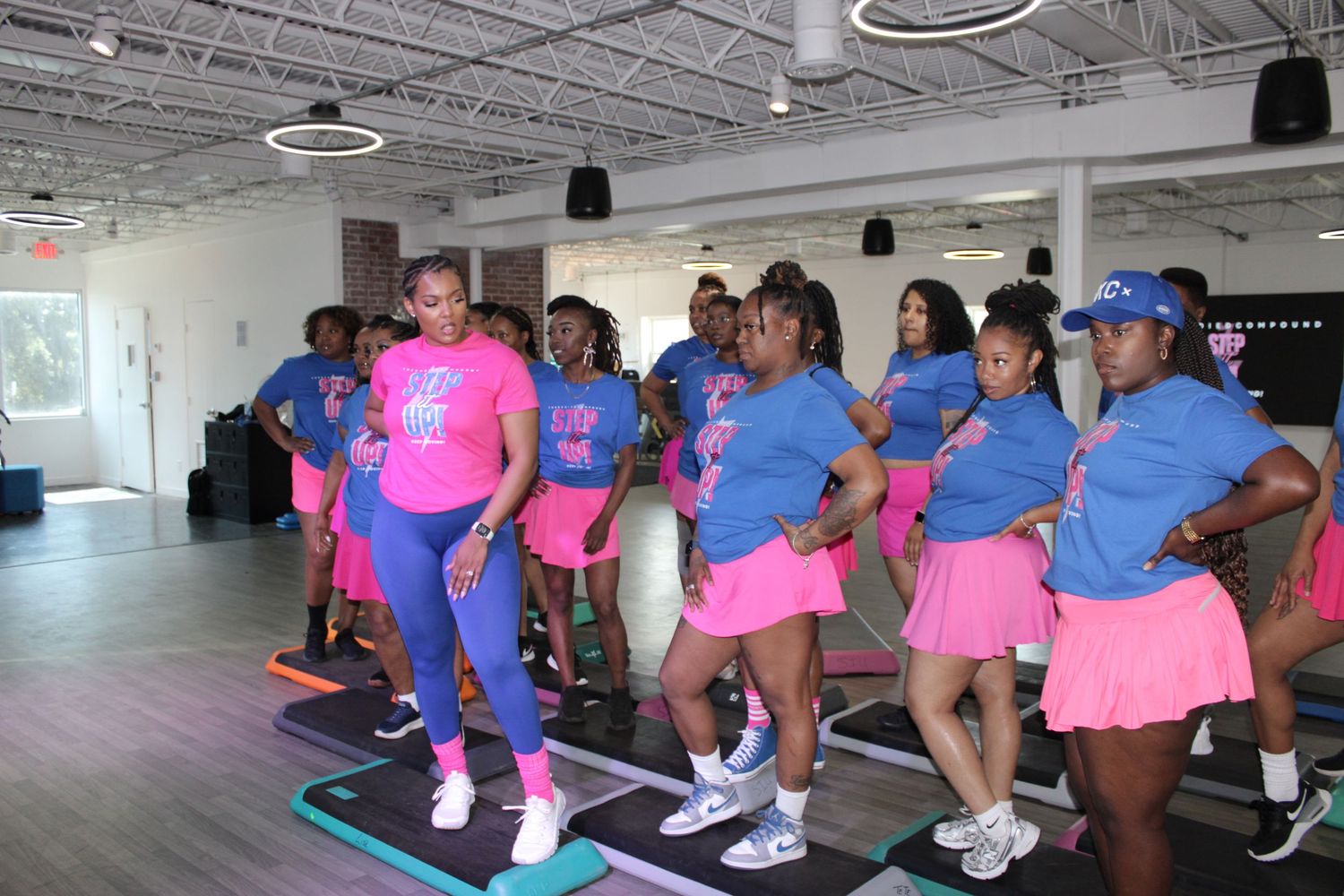 Women in matching pink and blue Step It Up outfits participate in a step aerobics class in a bright modern fitness studio.
